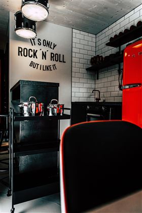 A modern kitchen with a red refrigerator and a stylish shelf.  
On the wall is the saying "It's only rock 'n' roll, but I like it."