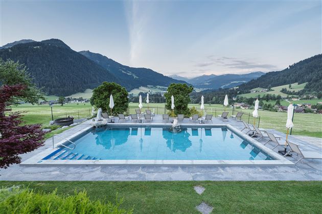 A beautiful pool with sun loungers, surrounded by green meadows and mountains. The sky is clear and shows soft colors of the sunset.