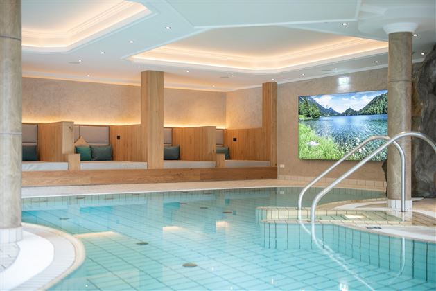 A relaxing wellness area with a swimming pool and cozy loungers. On the wall, there is a screen displaying a landscape image.