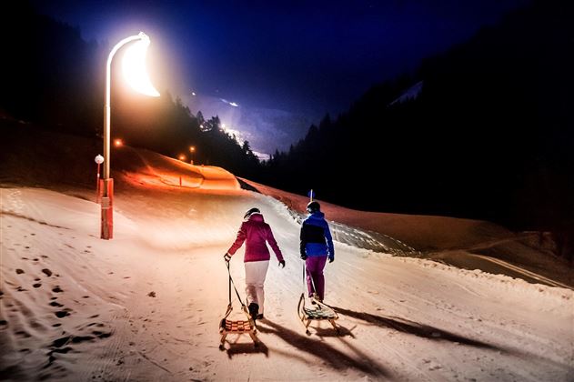 Two people are pulling sleds on a lit snow slope at night. In the background, the lights of the mountain are shining.