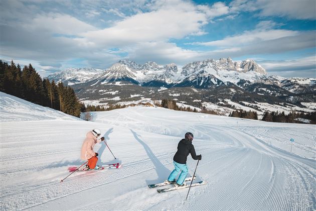 Two skiers are skiing on a snowy slope. In the background, impressive mountains and a blue sky are visible.