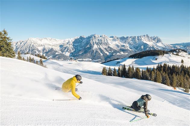 Two skiers are skiing over fresh snow in the mountains. In the background, majestic mountains and a clear blue sky can be seen.