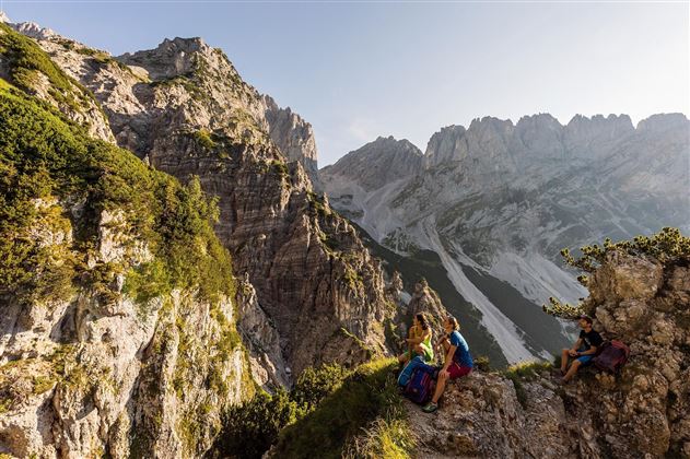 A stunning mountain landscape with steep rocks and green forests. Two hikers enjoy the view of the surrounding peaks.
