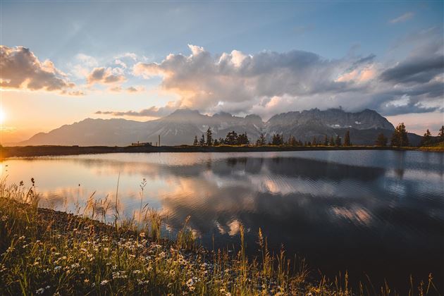A tranquil lake reflects the mountains and the sky at sunset. The edge is lined with green meadows and flowers.