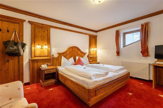 A cozy bedroom with a large double bed and wooden paneling. The room has red carpets and windows with decorative curtains.