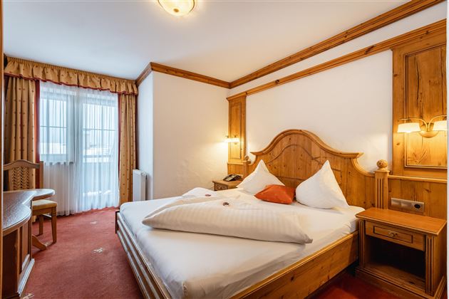 A cozy hotel room with wooden decor and a large bed. There is a table, a chair, and a window with curtains.
