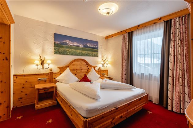 A cozy bedroom with a wooden bed and red carpet. The room is brightly lit and has large windows looking outside.