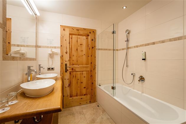 A modern bathroom with a wooden door and large mirrors. There is a bathtub, two sinks, and a shower.