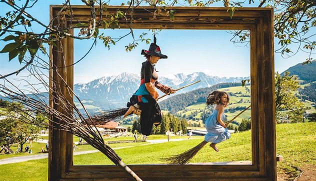 A girl and a witch are happily flying on brooms in a picturesque landscape frame. In the background, green hills and mountains are visible.