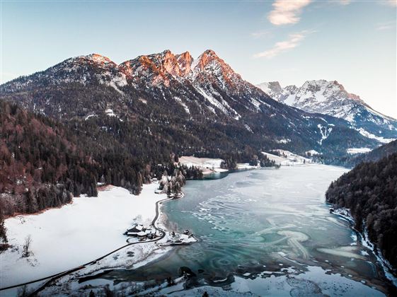 A charming landscape with snow-covered mountains and a frozen lake. The sky is clear and the colors of the sunset are visible.