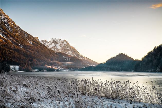 A winter landscape with a frozen lake and snow-covered mountains in the background. The sky is clear and the sun is shining gently.