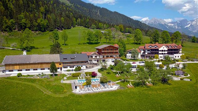 A picturesque landscape with a hotel complex, surrounded by forests and mountains. In the foreground, there is a pool area and green meadows.