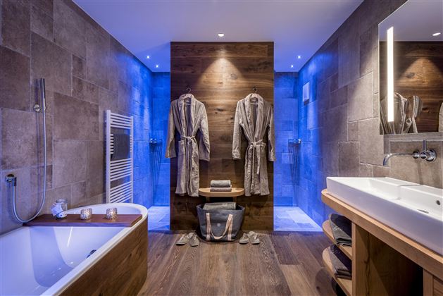 A modern bathroom with a bathtub and elegant wood paneling. Two bathrobes hang next to a sink with soft lighting.