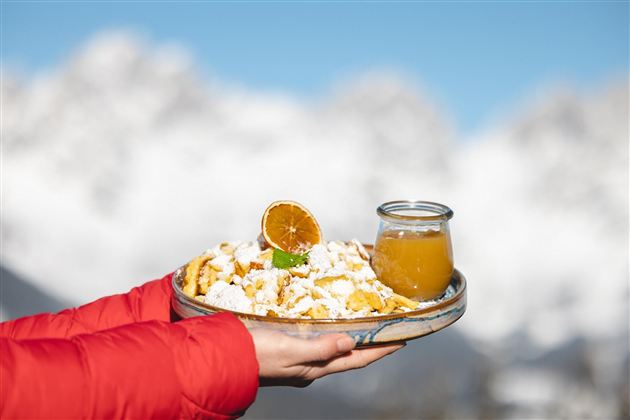 Ein Teller mit einer köstlichen Süßspeise und einem Glas Orangensaft wird von einer Hand gehalten. Im Hintergrund sind schneebedeckte Berge zu sehen.