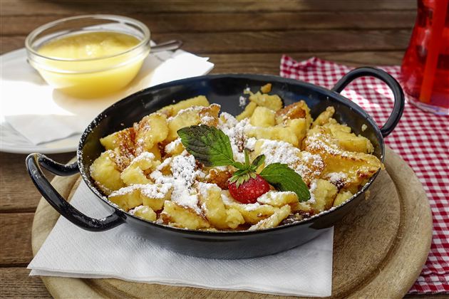 A delicious Kaiserschmarrn in a black pan, garnished with powdered sugar and a fresh mint leaf. Next to it is a bowl of applesauce.