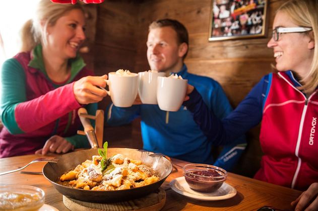Drei Personen stoßen mit Tassen an und genießen eine Mahlzeit in einer gemütlichen Hütte. Auf dem Tisch liegt eine Schüssel mit einem leckeren Dessert.