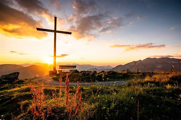 A cross silhouette against a stunning sunset over the mountains. Green meadows and flowers complement the peaceful scene.