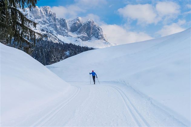 A snow-covered landscape with a skier on a ski trail. In the background, majestic mountains and a blue sky can be seen.