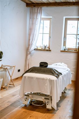 A quiet wellness room with a treatment table and large windows. The wooden floor and soft colors create a relaxed atmosphere.