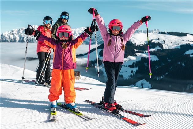 A group of skiers is standing on a snow-covered slope. Two excited children in colorful ski outfits are raising their hands in the air.