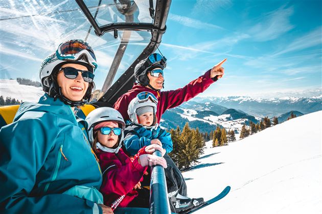 A family in a ski lift enjoying the view of the snow-covered mountains. They are wearing ski helmets and goggles and appear cheerful.
