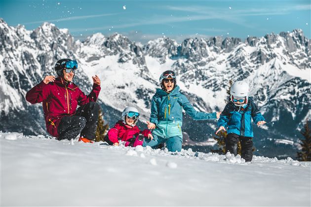 A family is playing in the snow with snow-covered mountains in the background. Everyone is wearing ski helmets and colorful winter clothing.