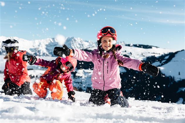 Three children are enthusiastically playing in the snow. They are laughing, throwing snowballs into the air, and wearing colorful ski clothing.