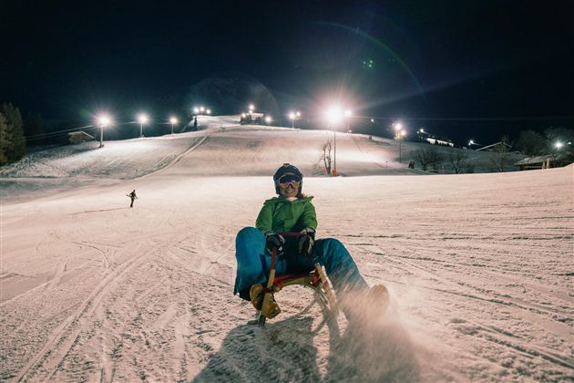 A nighttime sledding fun on a snowy slope. In the background, the lights of the ski slope shine.