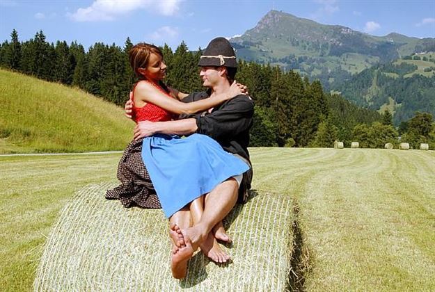 A couple is sitting on a hay bale in a beautiful, green landscape. The surroundings are hilly, with trees and a clear sky in the background.