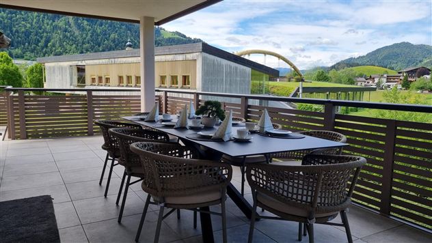 A cozy dining area on a terrace with a large table and chairs. In the background, green mountains and a blue sky can be seen.
