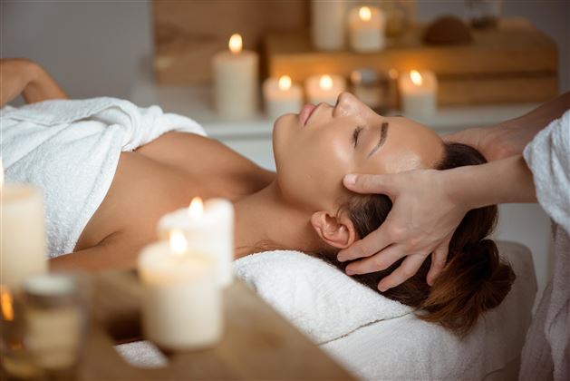 A relaxed woman is receiving a head massage on a couch. In the background, candles are burning, creating a calming atmosphere.