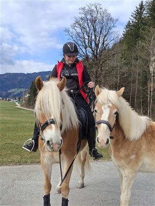 A woman is riding a horse and next to her stands another pony. The landscape is green and wooded, with a clear sky in the background.