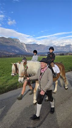 Two children are riding on ponies, accompanied by an older man. In the background, there is a green meadow and mountains under a blue sky.
