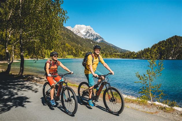 Two people are riding bicycles along a clear lake. In the background, there are mountains and blooming nature.