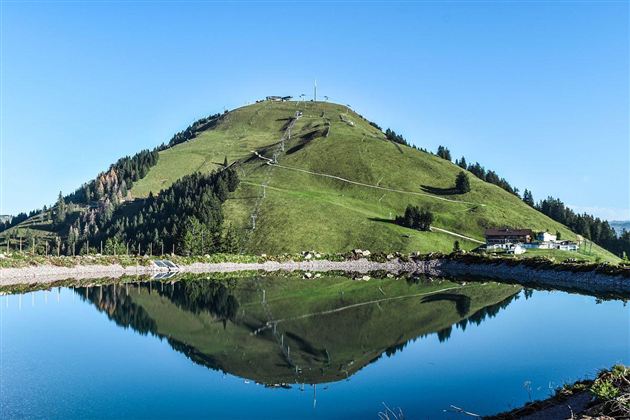 A beautiful mountain with green meadows and pine trees. The clear sky is reflected in the calm water of a pond.