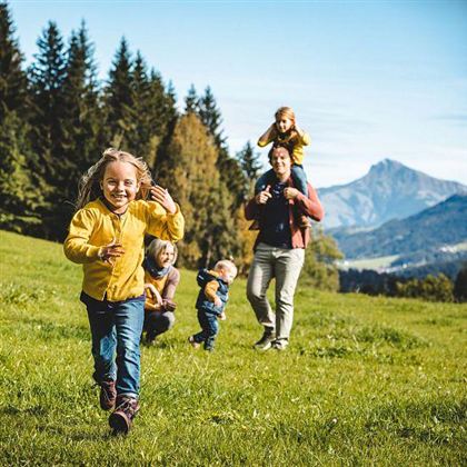 A cheerful family is playing in a green meadow with mountains in the background. Children are running and laughing while the adults accompany them.
