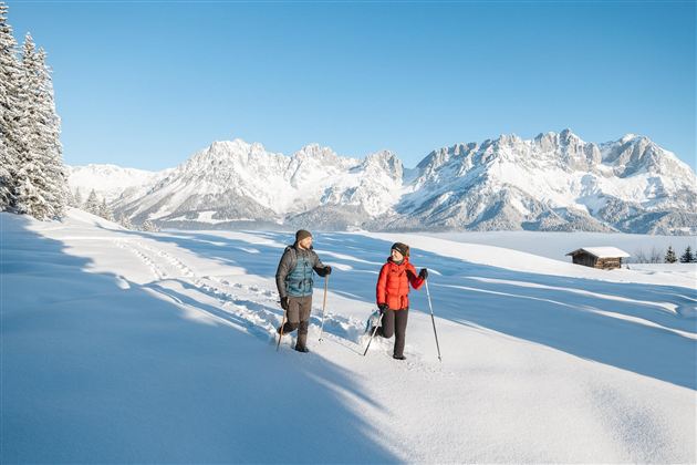 Two people are hiking in the snow with ski poles in front of an impressive mountain backdrop. The clear sky and the snowy landscape create a peaceful winter atmosphere.