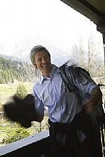 A cheerful man stands on a balcony holding a jacket in his hand. In the background, mountains and trees can be seen.