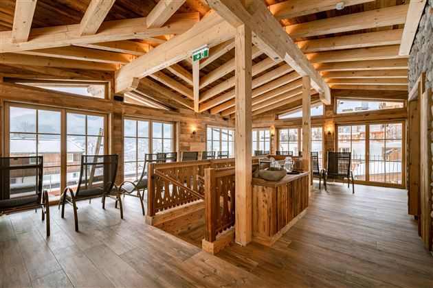 A cozy interior with a wooden ceiling and large windows. The room is furnished with comfortable chairs and natural light.