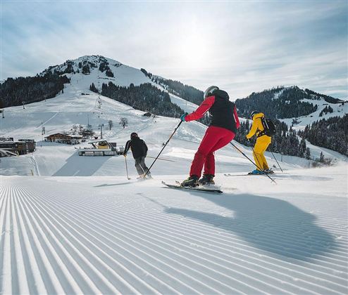 Drei Skifahrer fahren eine präparierte Piste hinunter. Im Hintergrund sind schneebedeckte Berge und ein Skilift zu sehen.