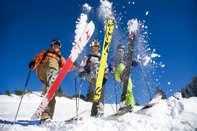 Three skiers are standing in the snow and showing their skis. The sky is clear and blue, and fresh snow is being stirred up.