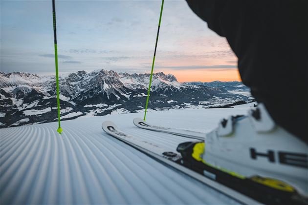Eine Skisicht auf schneebedeckte Berge im Hintergrund. Im Vordergrund sind Skier und ein Skischuh zu sehen.