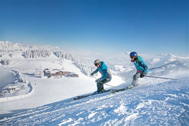 Two skiers are skiing down the snowy slope. In the background, there are snow-covered mountains and a blue sky.