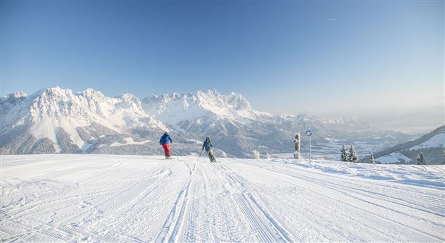 Two skiers are skiing on a snowy slope with impressive mountain peaks in the background. The sky is clear and sunny.