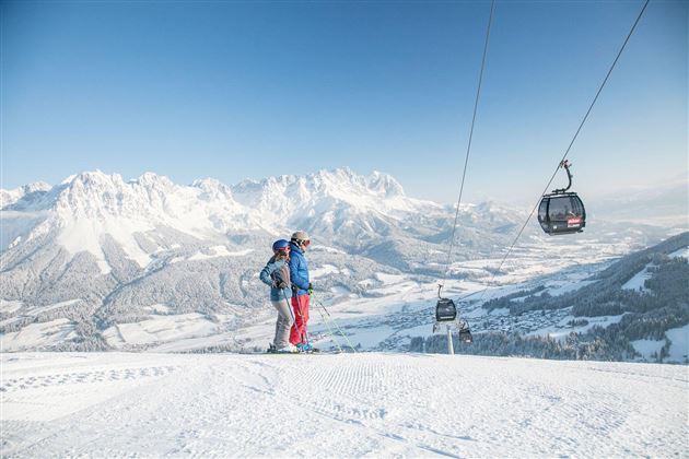 Ein Paar steht auf einem schneebedeckten Berggipfel mit Blick auf die winterliche Landschaft. Im Hintergrund sind die Berge und eine Seilbahn zu sehen.