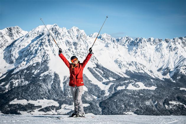 A person stands on a snow-covered mountain, lifting the ski poles into the air. In the background, impressive, snow-covered mountains can be seen.
