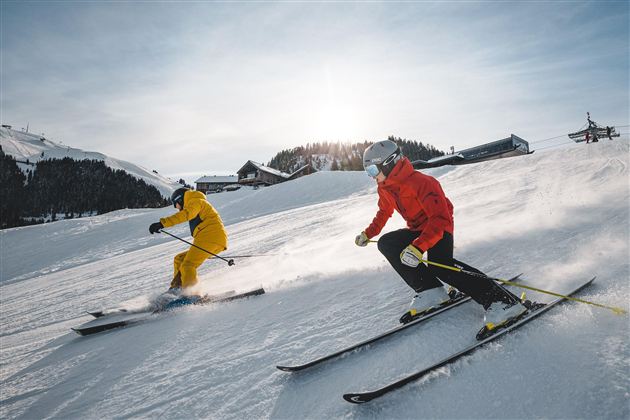 Two skiers are going down a snow-covered slope. One is wearing a yellow suit and the other a red one, while the sun shines in the background.