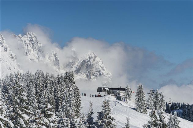 A snow-covered mountain landscape with tall fir trees and a ski lift. In the background, impressive mountains and blue sky can be seen.