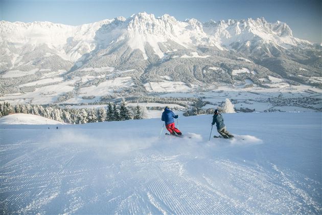 Two skiers glide over a snowy slope. In the background, majestic mountains stretch under a clear sky.