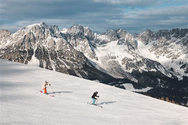 Zwei Skifahrer fahren eine verschneite Piste hinunter. Im Hintergrund sehen wir beeindruckende Berge unter einem bewölkten Himmel.